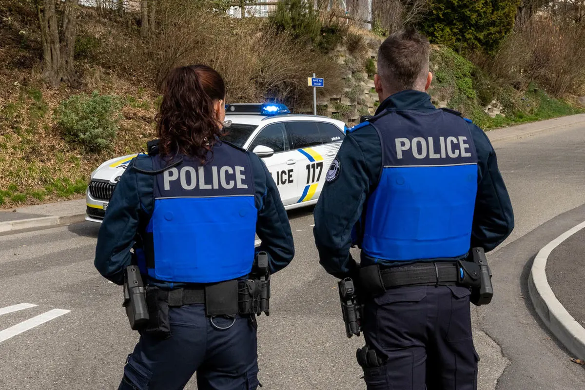 Zwei Polizisten in blauen Westen stehen mit dem Rücken zur Kamera vor einem Streifenwagen mit aktivem Blaulicht.