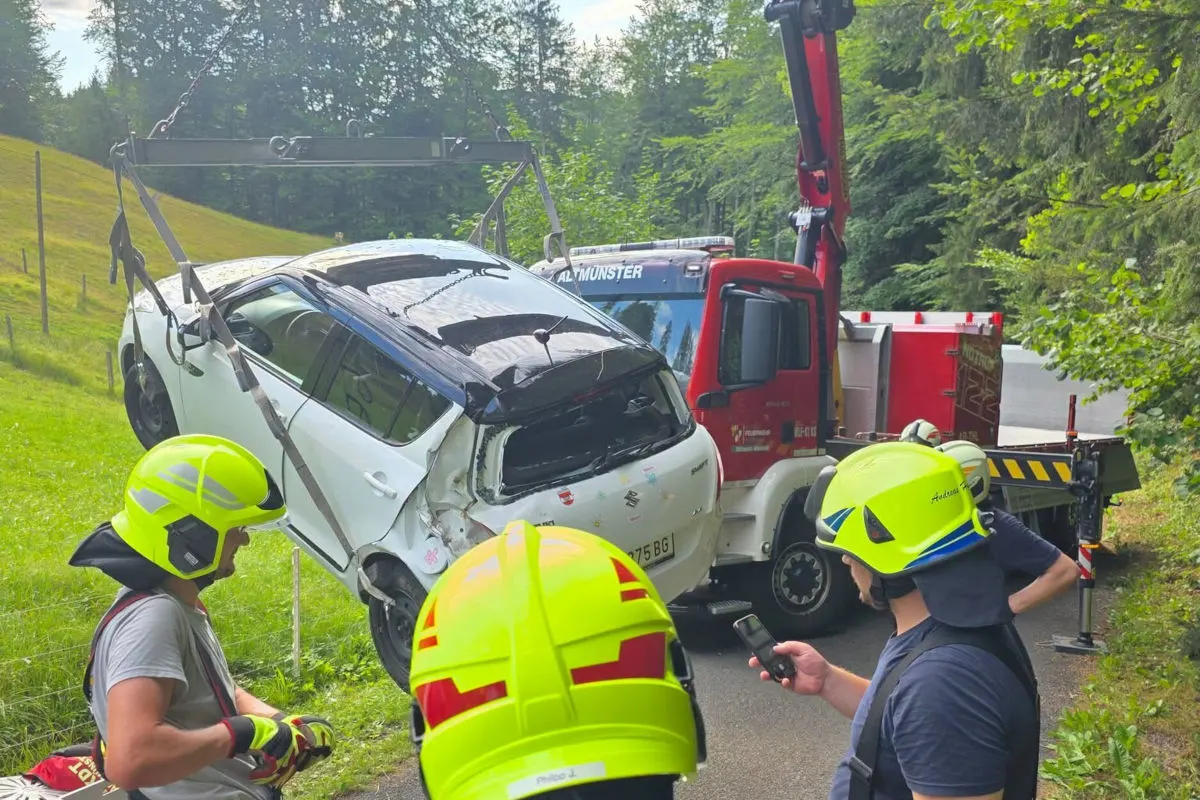 Ein stark beschädigtes weißes Auto wird von einem Fahrzeugkran gehoben, umgeben von Feuerwehrleuten mit gelben Helmen. Im Hintergrund sind grüne Bäume sichtbar.