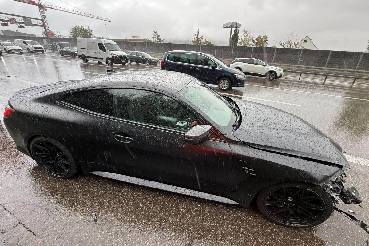 Ein schwarzer Sportwagen mit Frontschaden steht auf einer regennassen Straße. Im Hintergrund fahren weitere Autos vorbei.