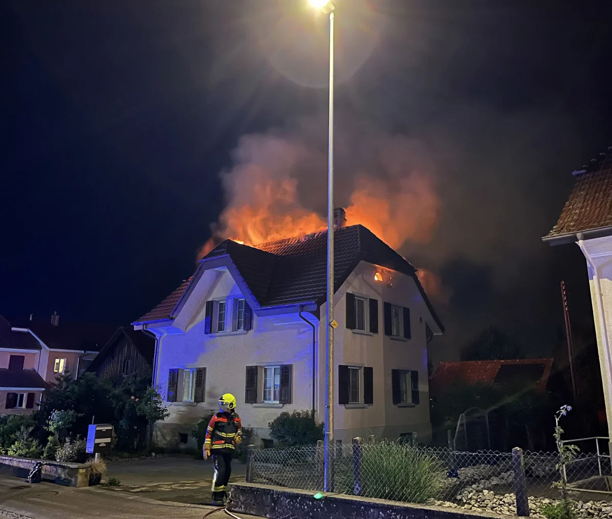 Das Bild zeigt ein brennendes Einfamilienhaus bei Nacht. Dichte Flammen und Rauch kommen aus dem Dach. Ein Feuerwehrmann steht im Vordergrund.
