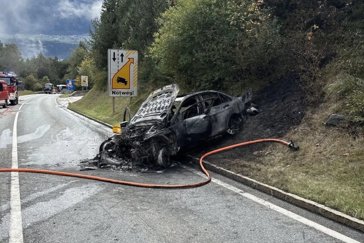 Ein ausgebrannter Pkw liegt neben einer Straße auf einem Notweg. Im Hintergrund sind Feuerwehrfahrzeuge sichtbar.