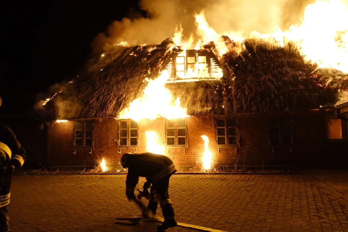 Ein reetgedecktes Gebäude ist in der Nacht vollständig in Flammen. Feuerwehrleute sind im Vordergrund zu sehen, die den Brand bekämpfen.