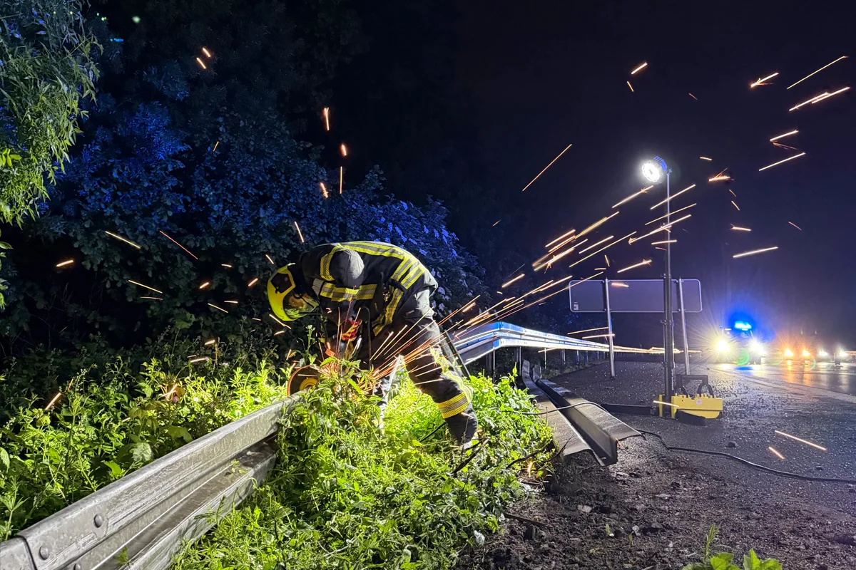 Ein Feuerwehrmann beugt sich über eine Leitplanke und bearbeitet sie mit einem Winkelschleifer, während Funken sprühen. Der Hintergrund zeigt eine dunkle, bewaldete Umgebung und ein beleuchtetes Einsatzfahrzeug.
