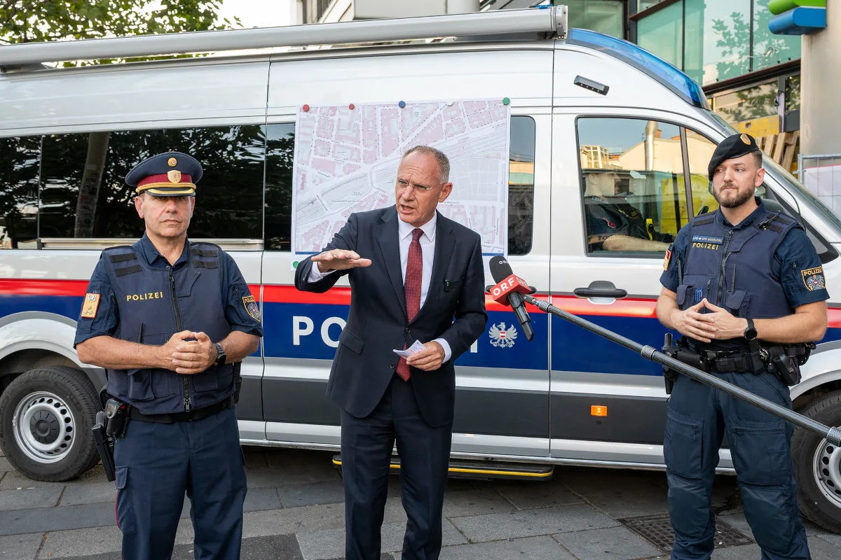 Ein Mann in Anzug spricht vor einem Polizeifahrzeug mit zwei uniformierten Polizisten daneben. Im Hintergrund ist ein Kartenausschnitt auf dem Fahrzeug montiert.