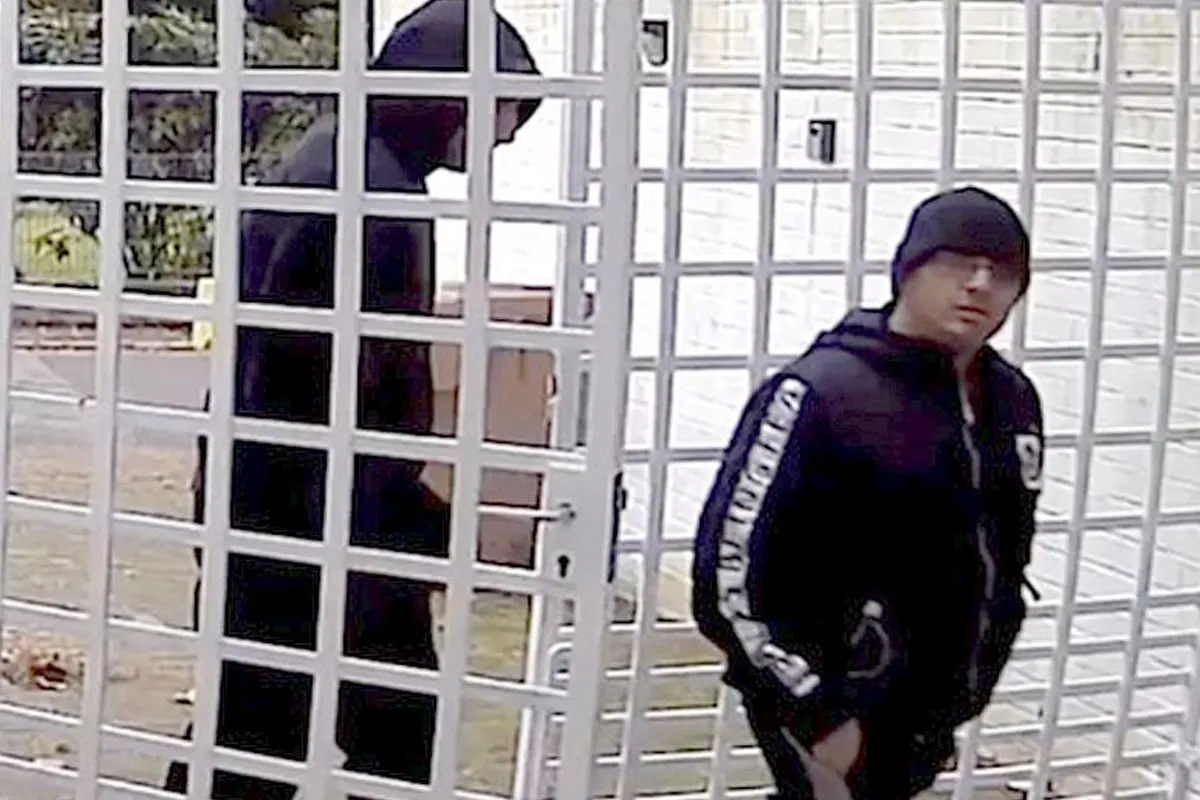 Zwei Personen in dunkler Kleidung mit Kapuzen und Mützen sind hinter einem Gittertor sichtbar. Eine Person blickt direkt zur Kamera.