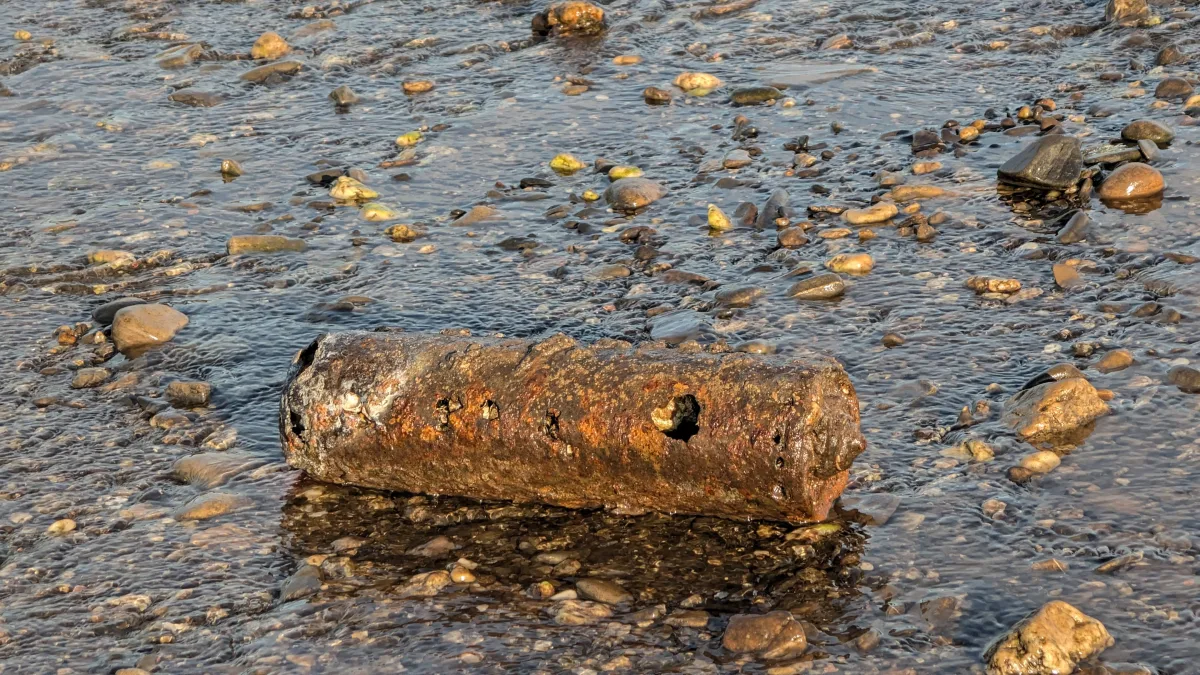 Eine rostige Metallröhre liegt in einem kiesbedeckten Flussbett. Die Röhre zeigt deutliche Abnutzungserscheinungen und liegt horizontal im flachen Wasser.