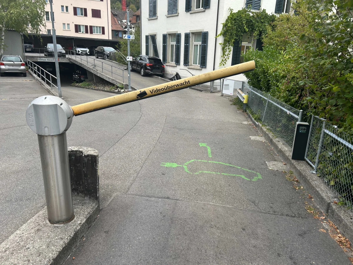 Eine Parkplatzeinfahrt mit einer Schranke im Vordergrund. Auf dem Boden sind grüne Markierungen sichtbar.