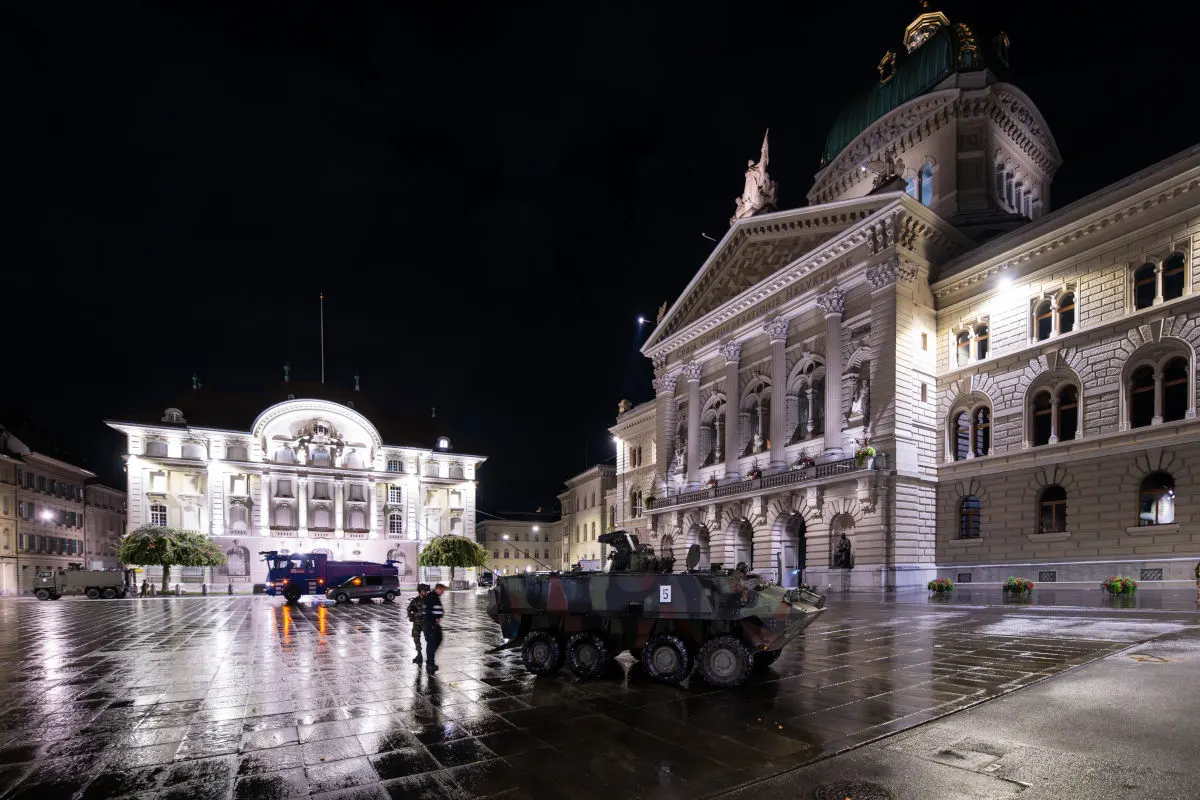 Vor dem beleuchteten Bundeshaus in Bern steht ein gepanzertes Fahrzeug auf einem nassen Platz. Zwei Personen in Uniform sind sichtbar.