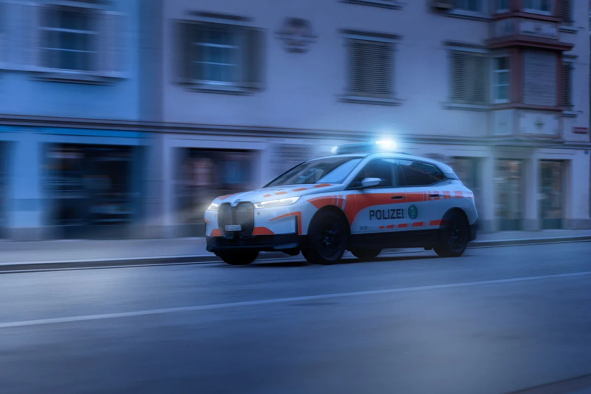 Ein Polizeiauto mit eingeschaltetem Blaulicht fährt schnell entlang einer Reihe von Gebäuden in der Dämmerung oder Nacht. Es handelt sich um ein modernes, weiß-orange markiertes Fahrzeug der Polizei.