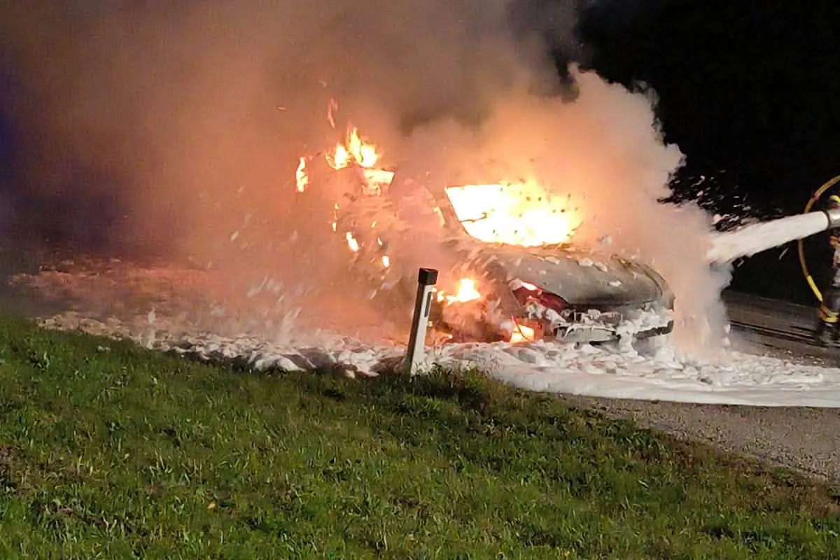 Ein Auto steht in Flammen und wird mit Schaum gelöscht, während Rauch in die Luft steigt. Daneben ist Wiese sichtbar.