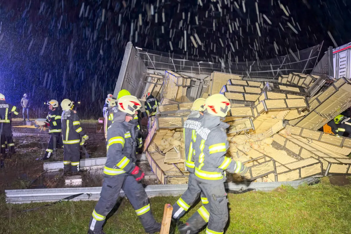Mehrere Feuerwehrleute in Schutzkleidung stehen vor einem umgestürzten Lkw, aus dem zahlreiche Kisten gefallen sind. Es regnet und es ist Nacht.