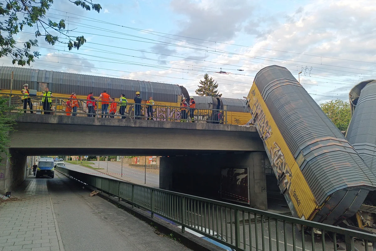 Auf einer Brücke sind entgleiste Güterwaggons zu sehen, daneben stehen Menschen in Warnwesten. Die Brücke überquert eine Straße.