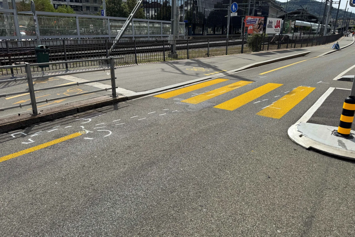 Eine Straße mit gelben Zebrastreifen und weißen Pfeilmarkierungen vor einem Bahnhof. Im Hintergrund sind Schienen und Gebäude zu sehen.