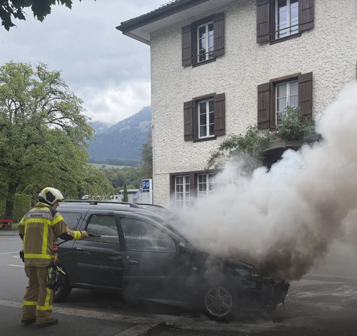 Ein Feuerwehrmann in Schutzkleidung ist dabei, ein stark rauchendes Auto vor einem Wohnhaus zu löschen. Im Hintergrund sind Bäume und Berge zu sehen.
