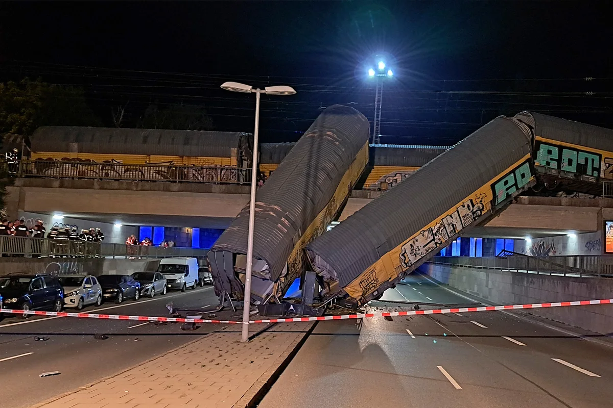 Zwei entgleiste Güterwagons hängen von einer Brücke und blockieren die Straße darunter, umgeben von Absperrband.