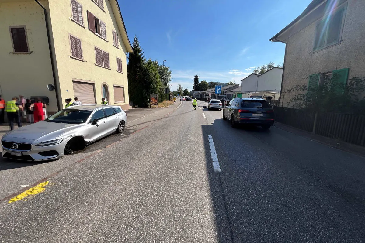 Ein silbernes Auto mit eingedrücktem Reifen steht am Straßenrand einer Wohngegend. Mehrere Personen in Westen und Autos sind im Hintergrund sichtbar.