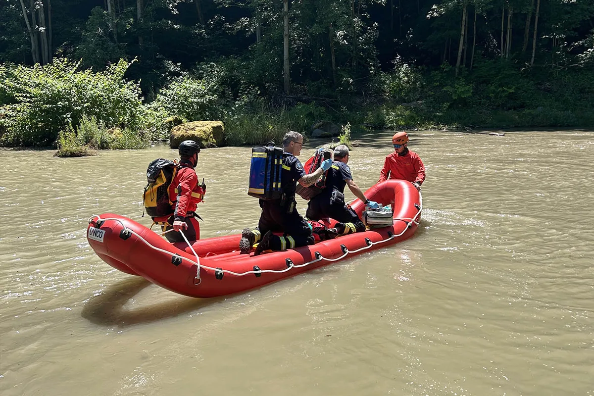 Mehrere Rettungskräfte befinden sich in einem roten Schlauchboot auf einem Fluss. Die Umgebung ist von bewaldeten Ufern gesäumt.