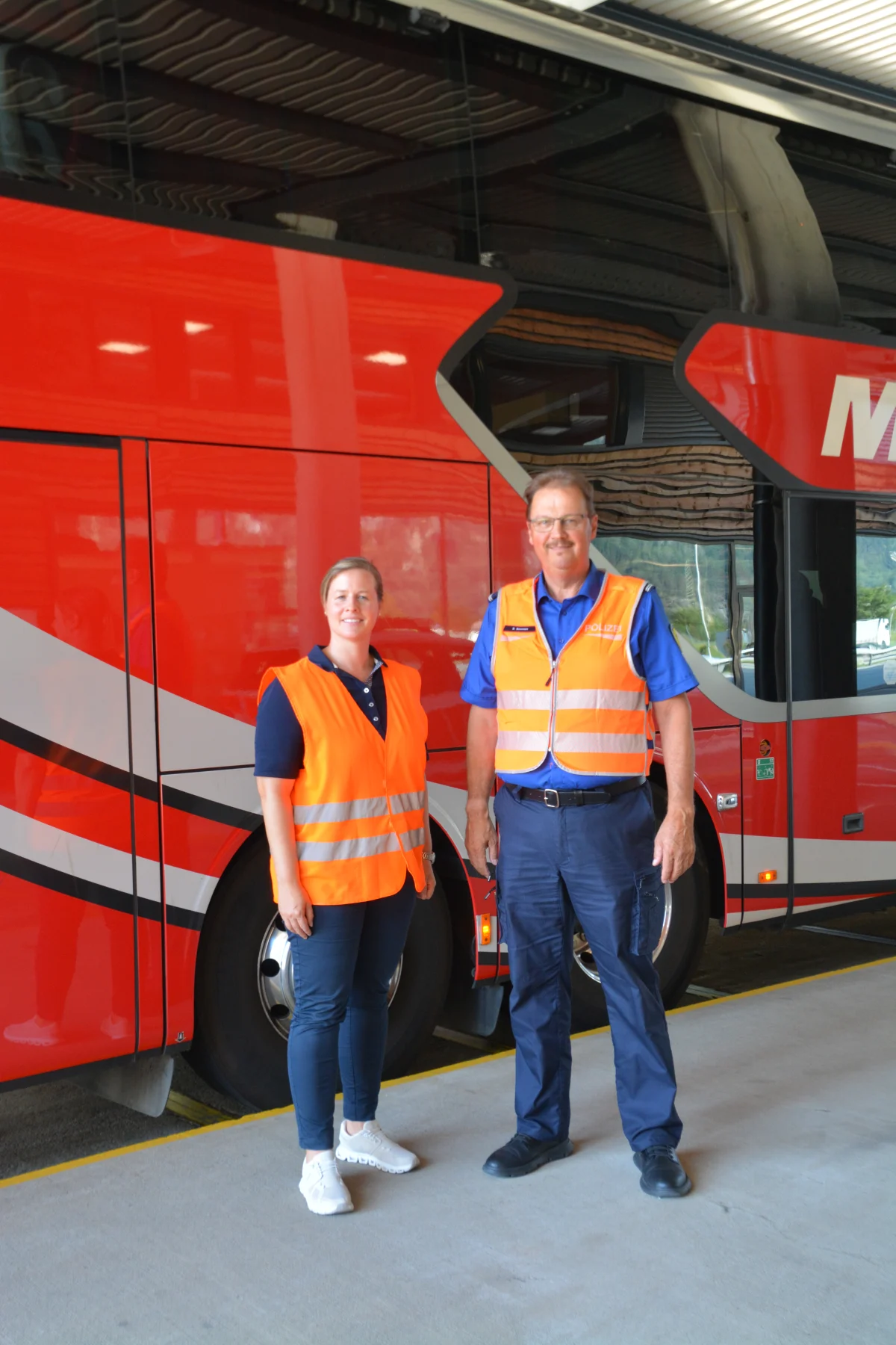 Zwei Personen stehen in orangefarbenen Warnwesten vor einem großen roten Reisebus in einer überdachten Umgebung.
