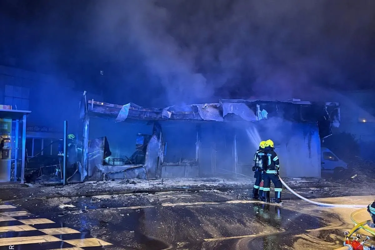 Zwei Feuerwehrleute mit gelben Helmen stehen vor einem völlig zerstörten Gebäude und löschen einen Brand bei Nacht. Rauch steigt in den Himmel.