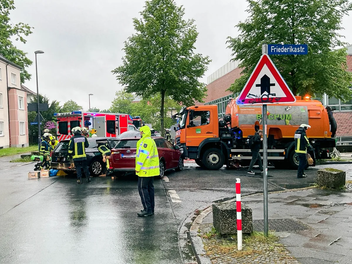 Ein Verkehrsunfall auf einer Stadtstraße mit mehreren Feuerwehrleuten in gelben Jacken, einem roten Rettungsdienstfahrzeug und einem orangefarbenen LKW mit 'Bochum'-Aufschrift.