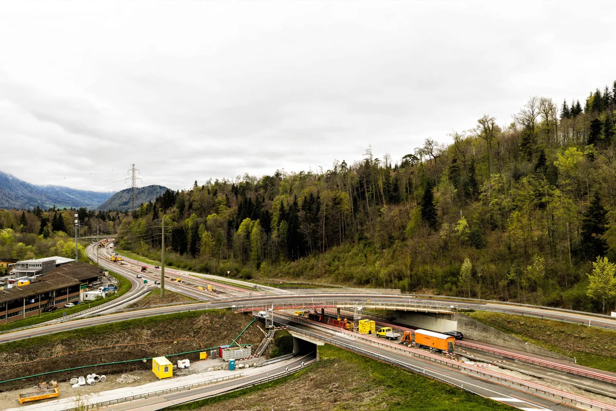Eine Straßenbaustelle auf einer kurvigen Strecke in einem bewaldeten Gebiet. Baustellenfahrzeuge und Absperrungen sind zu sehen.