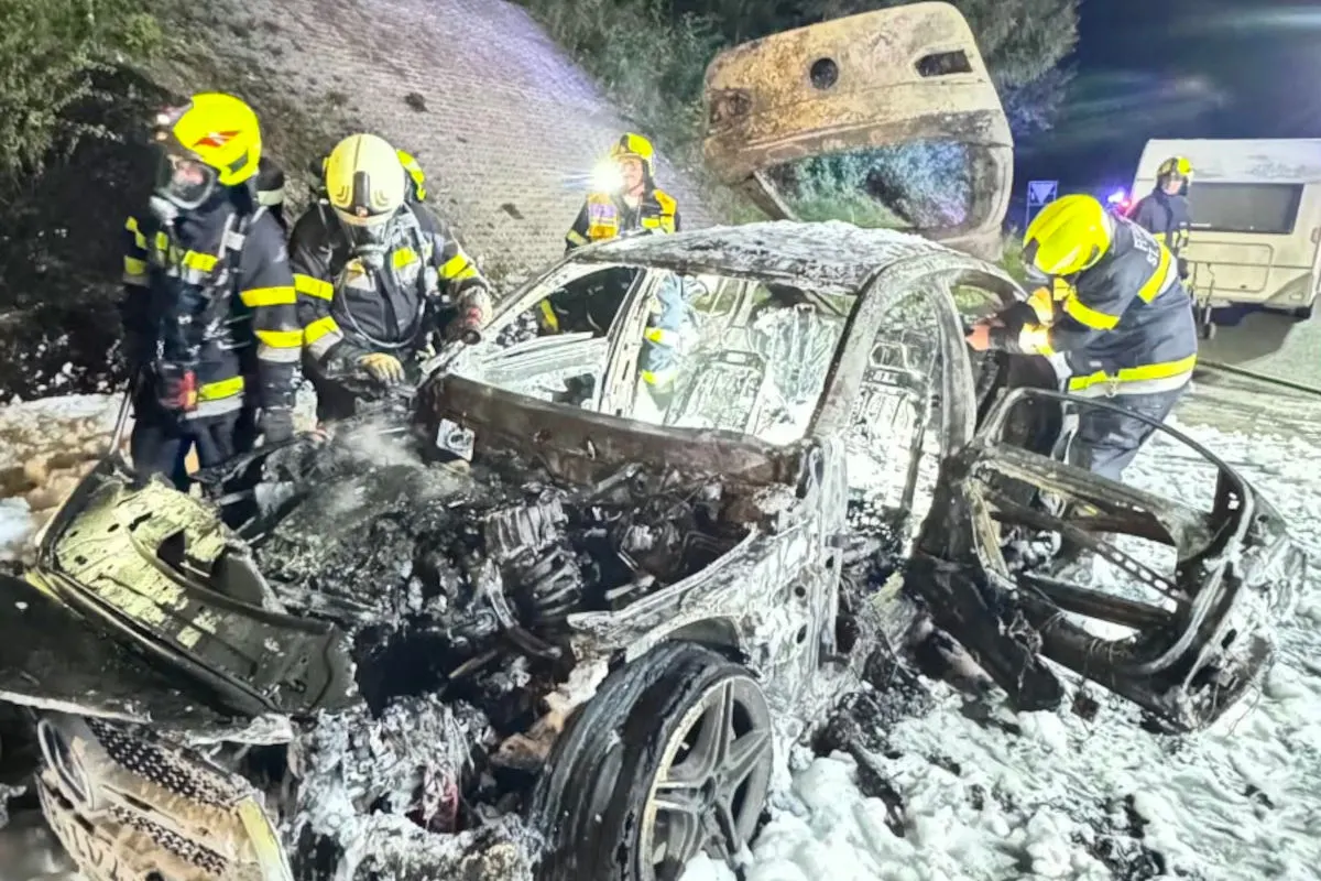 Mehrere Feuerwehrleute in Schutzkleidung arbeiten an einem stark beschädigten und verbrannten Fahrzeug. Es ist Nacht und der Unfallort ist mit Schaum bedeckt.