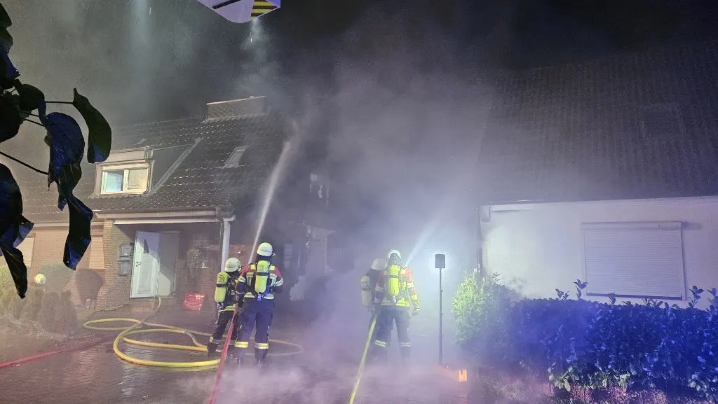 Drei Feuerwehrleute in Schutzkleidung bekämpfen bei Nacht einen Brand in einem Wohngebiet. Dicker Rauch zieht durch die Luft, während sie mit Schläuchen Wasser auf das Gebäude spritzen.