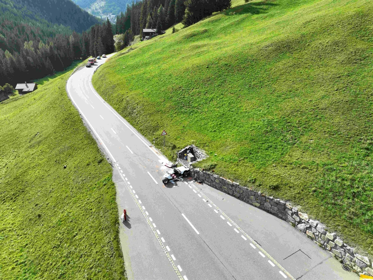 Ein Auto ist auf einer kurvigen Bergstraße gegen eine Mauer geprallt. Das Fahrzeug ist stark beschädigt, Rettungsfahrzeuge sind anwesend.