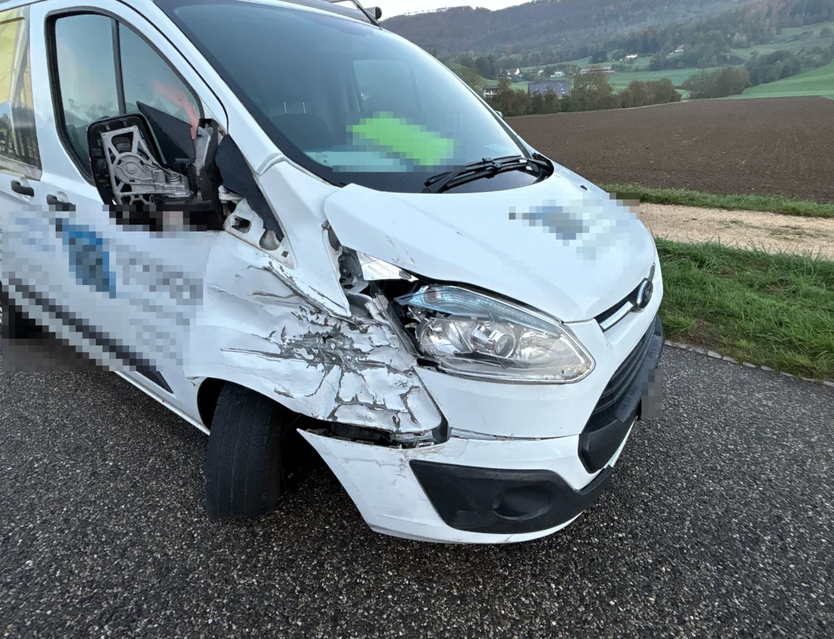 Ein weißer Transporter mit erheblichen Frontschäden steht auf einer asphaltierten Landstraße. Im Hintergrund sind Felder und Hügel sichtbar.
