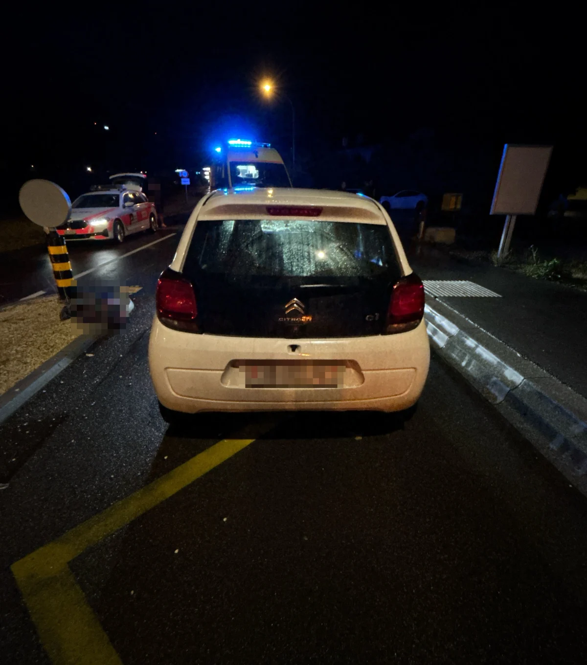 Ein weiß-schwarzer Citroën C1 parkt nachts auf einer nassen Straße, im Hintergrund leuchtet ein Fahrzeug mit Blaulicht.