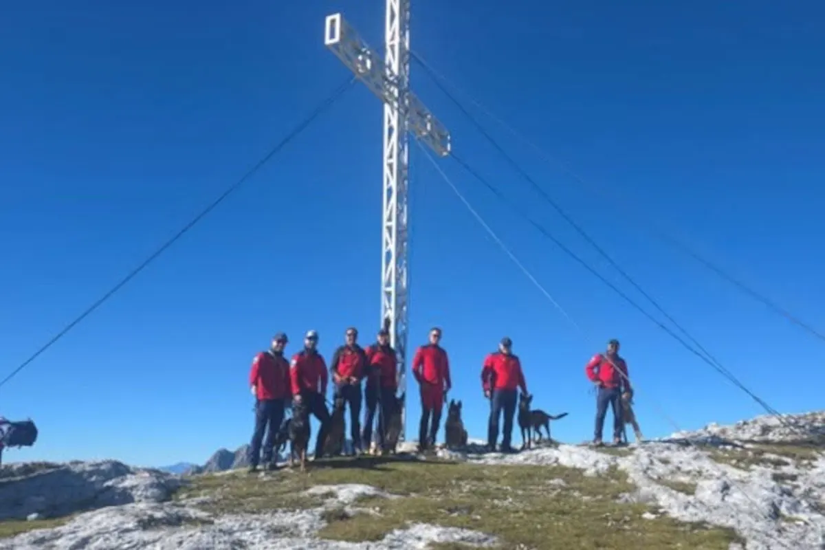 Eine Gruppe von Rettungskräften in roten Uniformen steht zusammen mit mehreren Hunden vor einem großen Gipfelkreuz. Der Himmel ist klar und blau.