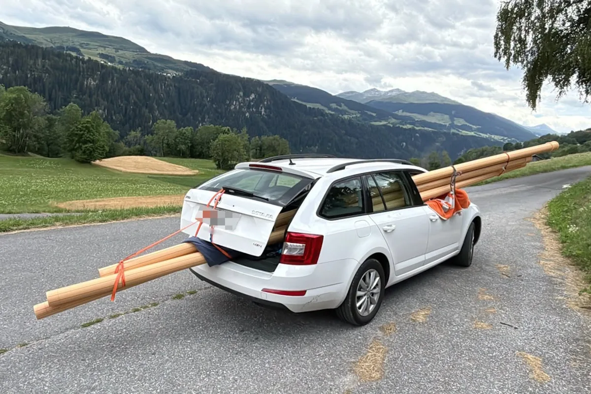 Ein weißer Kombi steht auf einer ländlichen Straße mit Holzstäben, die aus dem Kofferraum herausragen. Die Umgebung zeigt grüne Felder und Hügel im Hintergrund.