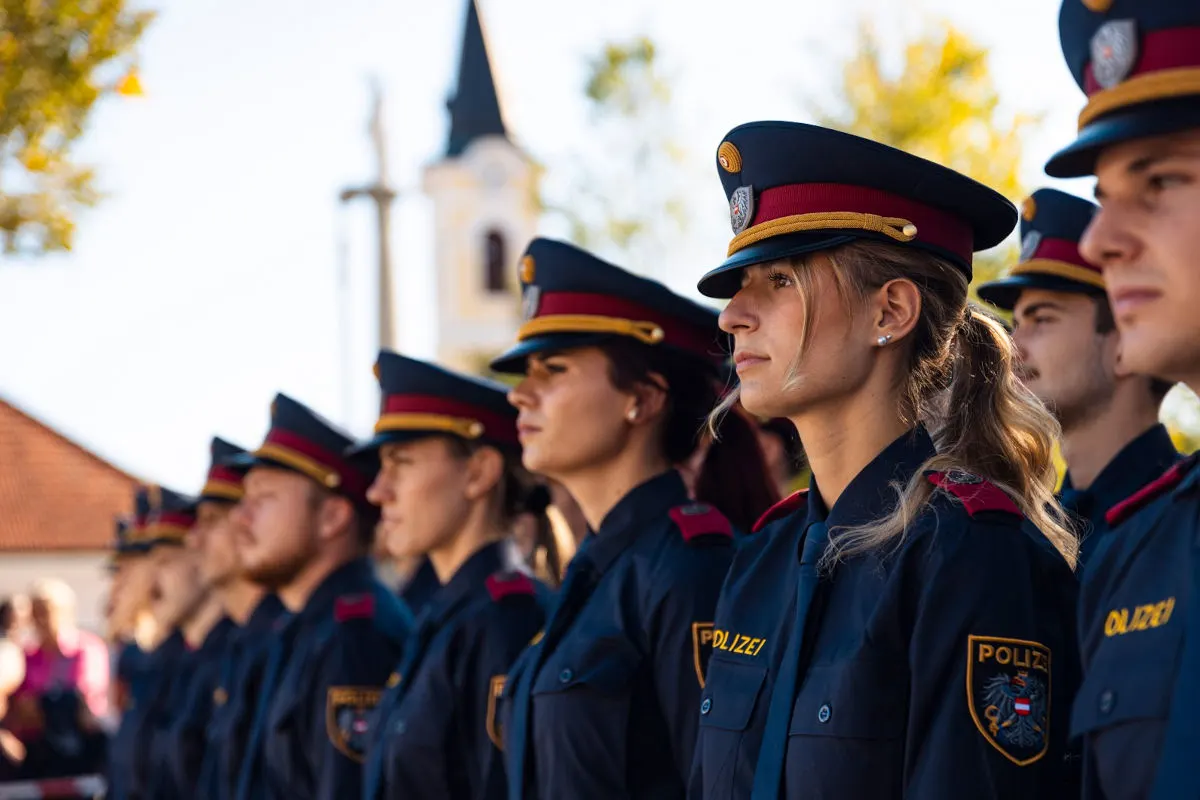 Eine Gruppe junger Polizisten in österreichischen Uniformen steht in einer Reihe, mit einer Kirche im Hintergrund.