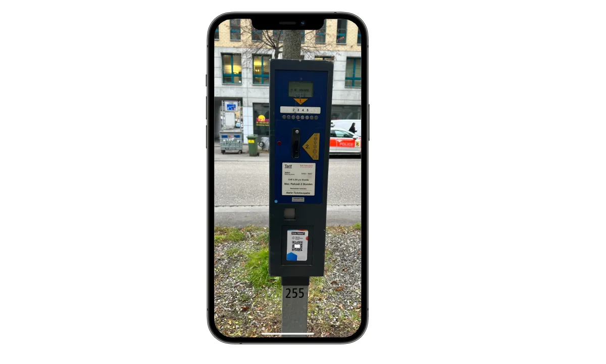 Im Vordergrund steht ein Parkscheinautomat, während im Hintergrund ein Fahrzeug der Polizei am Straßenrand parkt.