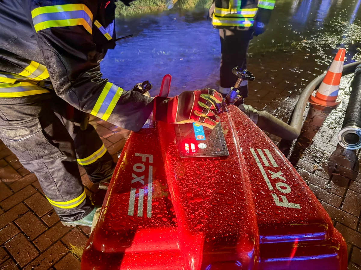 Ein Feuerwehrmann in Regenjacke und mit reflektierenden Streifen bedient bei Nacht eine nasse, rote Pumpe. Wasserleitungen und ein Verkehrskegel sind sichtbar.