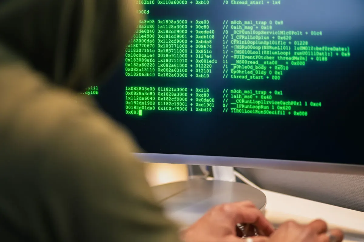 Eine Person sitzt vor einem Computer, auf dessen Bildschirm grüner Code zu sehen ist. Die Person trägt einen Kapuzenpulli.