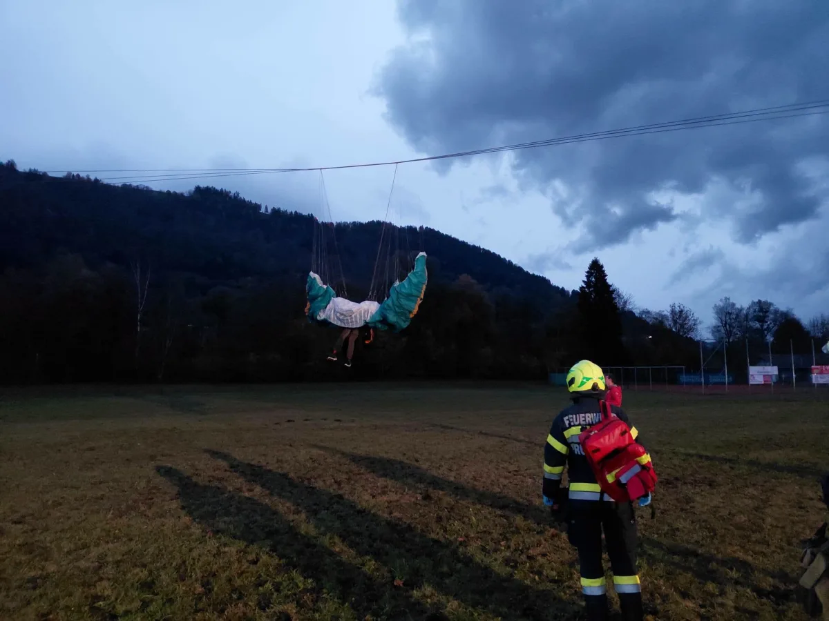 Ein Paraglider hängt in einer Stromleitung. Ein Feuerwehrmann mit Ausrüstung beobachtet die Szene im Freien bei wolkigem Wetter.