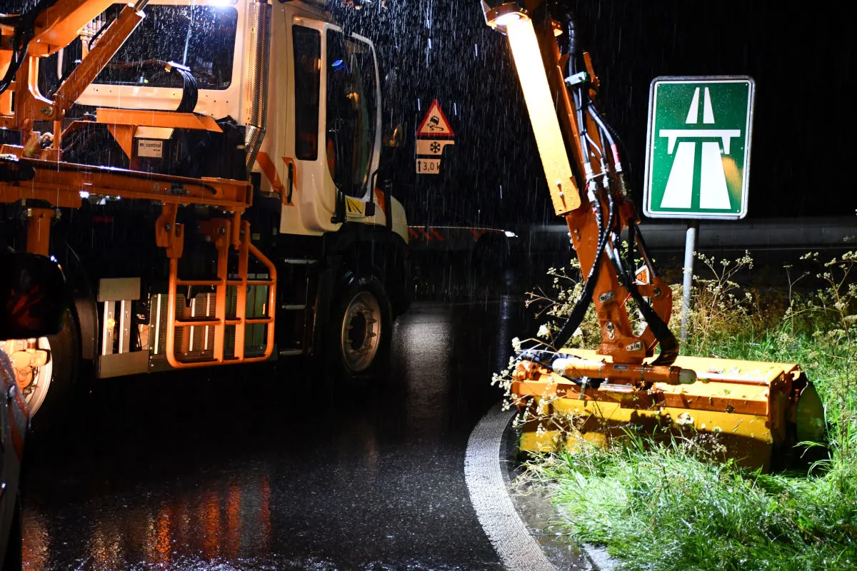Ein orangefarbener Wartungslastwagen mit Schneidegerät steht bei Nacht im Regen neben einem Autobahnschild. Das Fahrzeug reflektiert das Licht auf der nassen Straße.