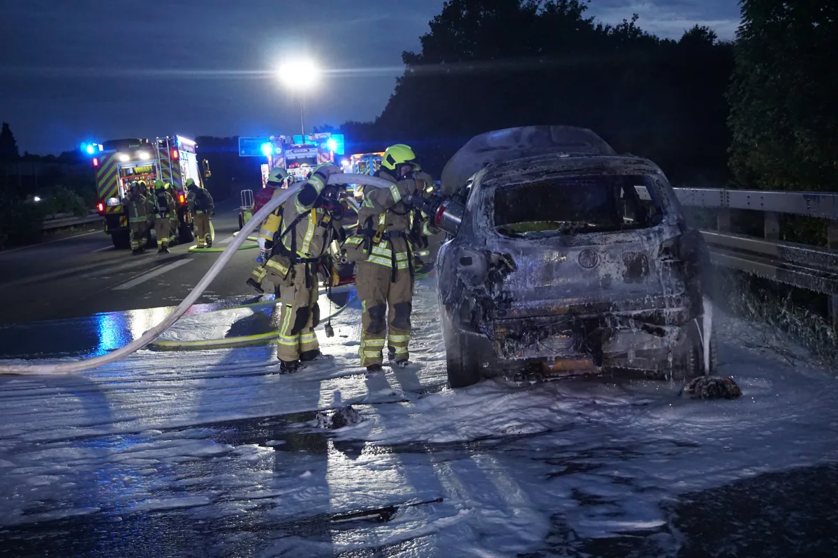 Zwei Feuerwehrleute stehen neben einem ausgebrannten Auto und löschen dieses mit Schaum. Im Hintergrund sind mehrere Einsatzfahrzeuge mit eingeschaltetem Blaulicht zu sehen.