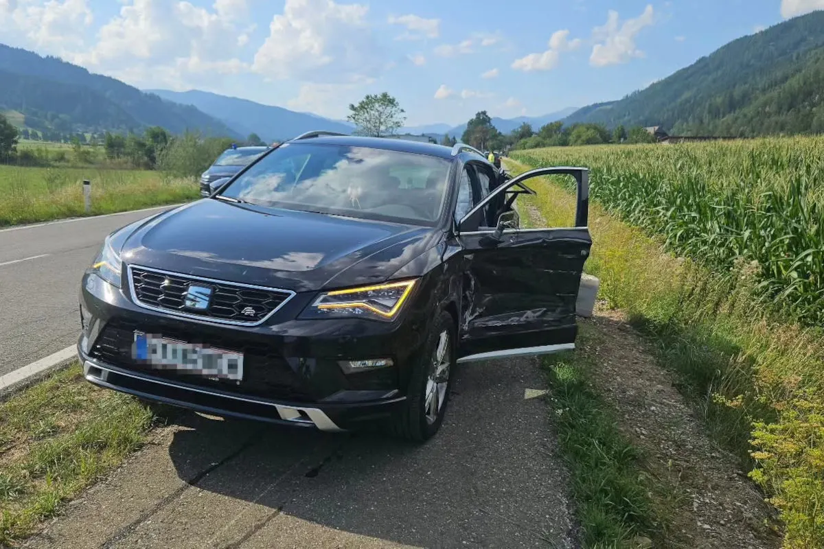 Ein schwarzer SEAT mit Seitenschäden steht am Straßenrand, die Fahrertür ist geöffnet. Die Umgebung ist ländlich mit Feldern und Hügeln im Hintergrund.