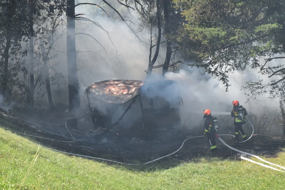 Zwei Feuerwehrleute in Schutzkleidung versuchen, einen rauchenden Anhänger im Wald zu löschen. Rauch quillt aus dem Anhänger zwischen den Bäumen.