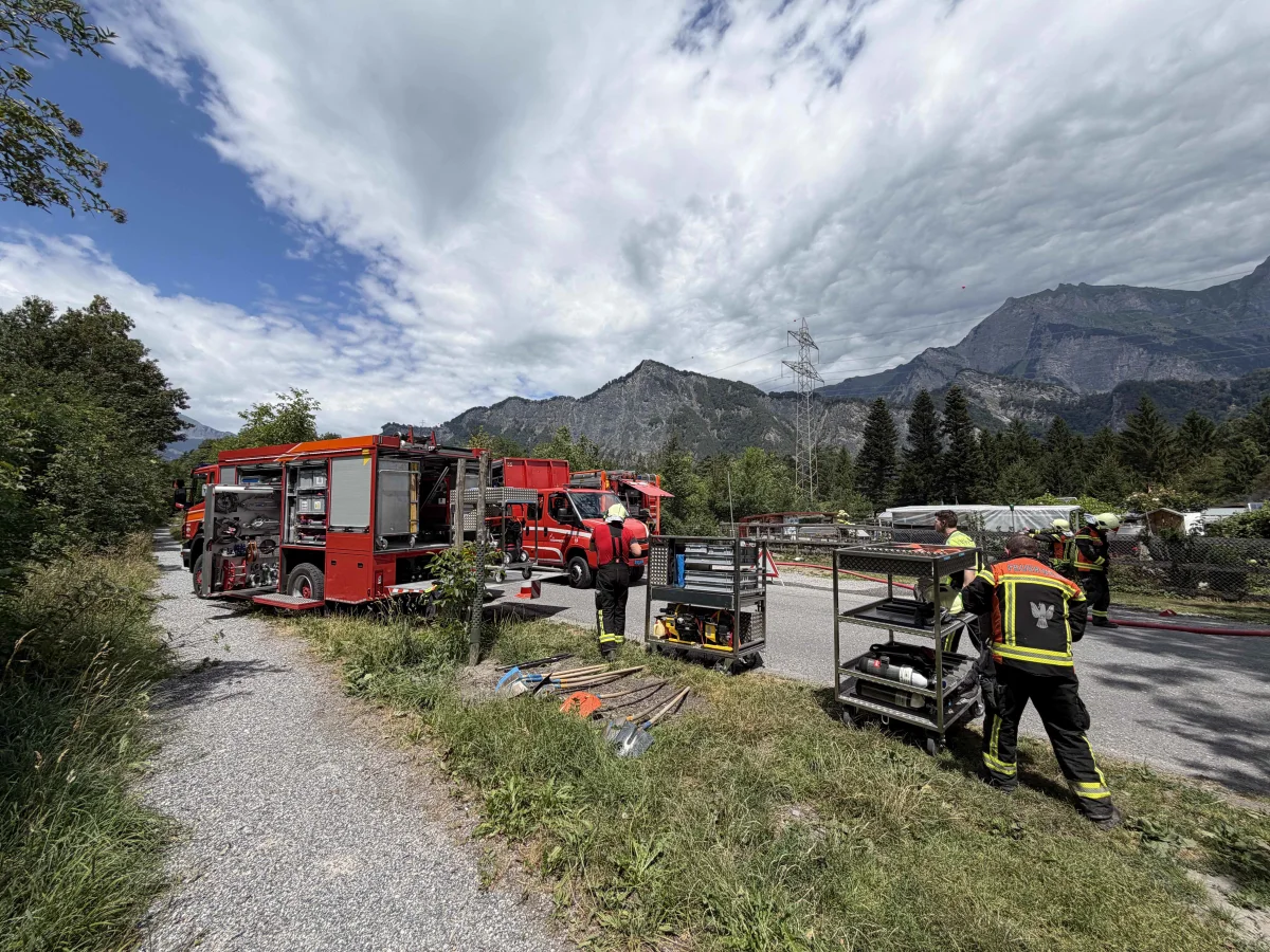 Mehrere Feuerwehrfahrzeuge stehen nahe einer Straße in einer alpinen Landschaft. Einsatzkräfte sind bei der Arbeit, umgeben von Bergen und Bäumen.