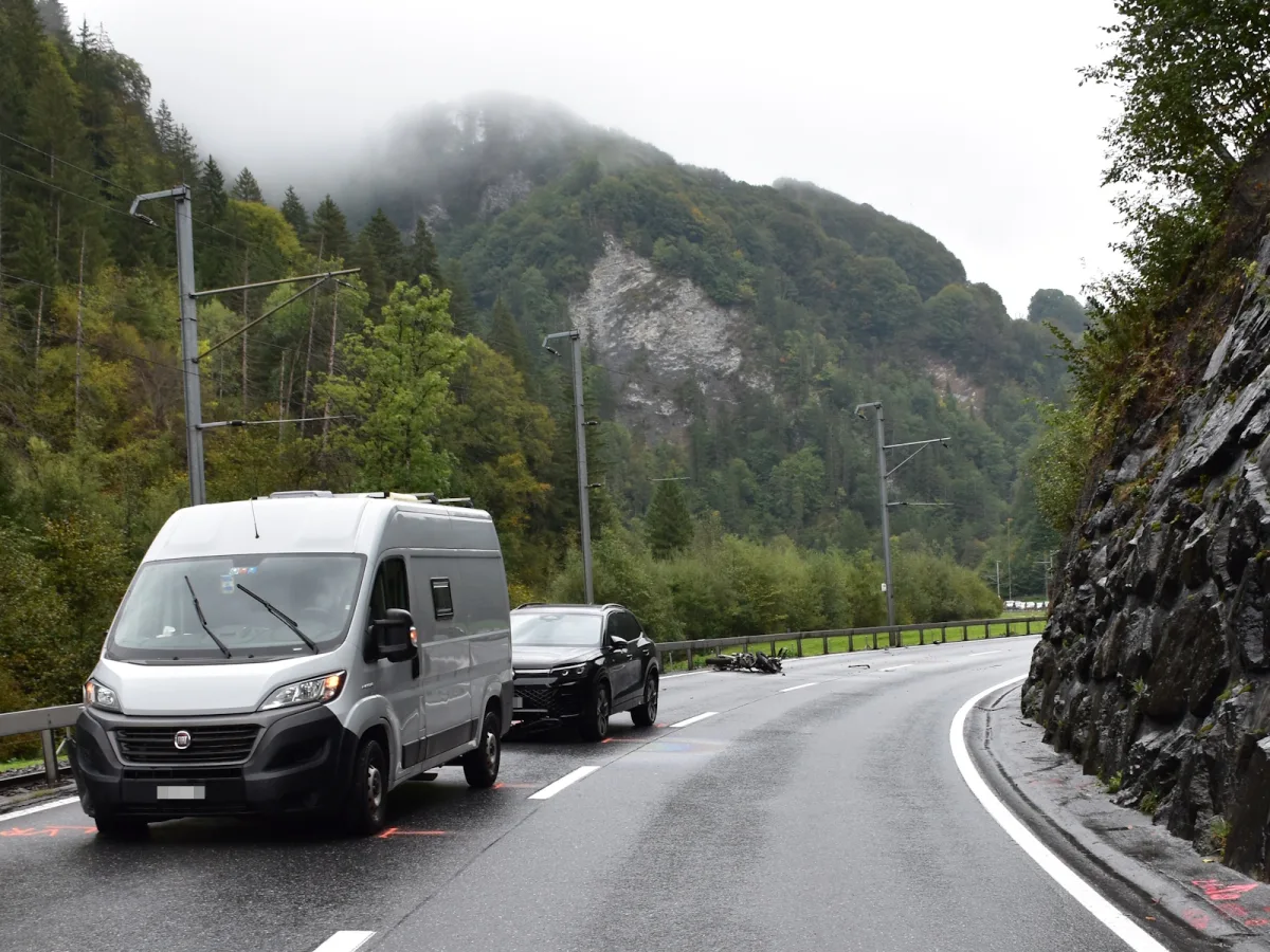 Ein silberner Van und ein schwarzes Auto stehen auf einer kurvigen, nassen Bergstraße. Im Hintergrund sind nebelbedeckte Berge und Wälder sichtbar.