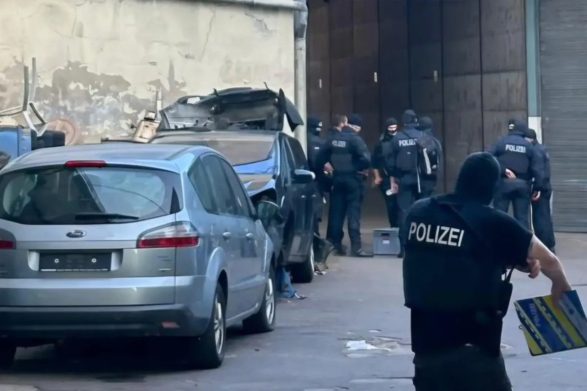 Eine Gruppe von Polizisten in Schutzkleidung betritt eine Lagerhalle. Im Vordergrund stehen zwei Autos ohne Kennzeichen.