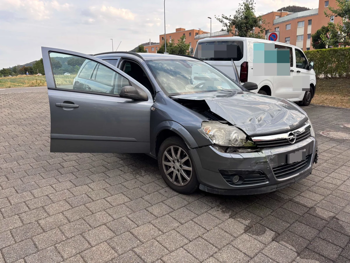 Ein grauer Opel, stark beschädigt an der Front, steht mit geöffneter Fahrertür auf einem gepflasterten Parkplatz neben einem weißen Van.