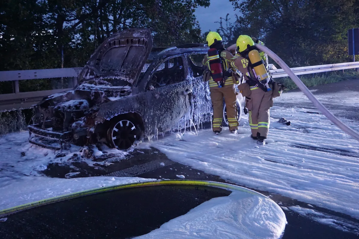 Zwei Feuerwehrleute in Schutzkleidung sind dabei, mit Schaum ein stark beschädigtes Auto zu löschen. Der Wagen ist völlig ausgebrannt.
