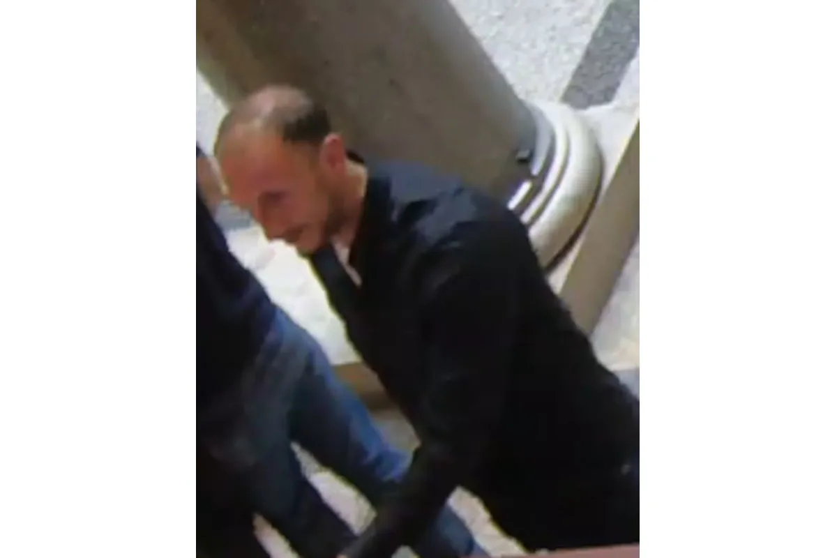 Ein Mann in dunkler Kleidung steht neben einer Säule in einem mit Steinen ausgelegten Innenraum. Die Szene wirkt öffentlich und belebt.