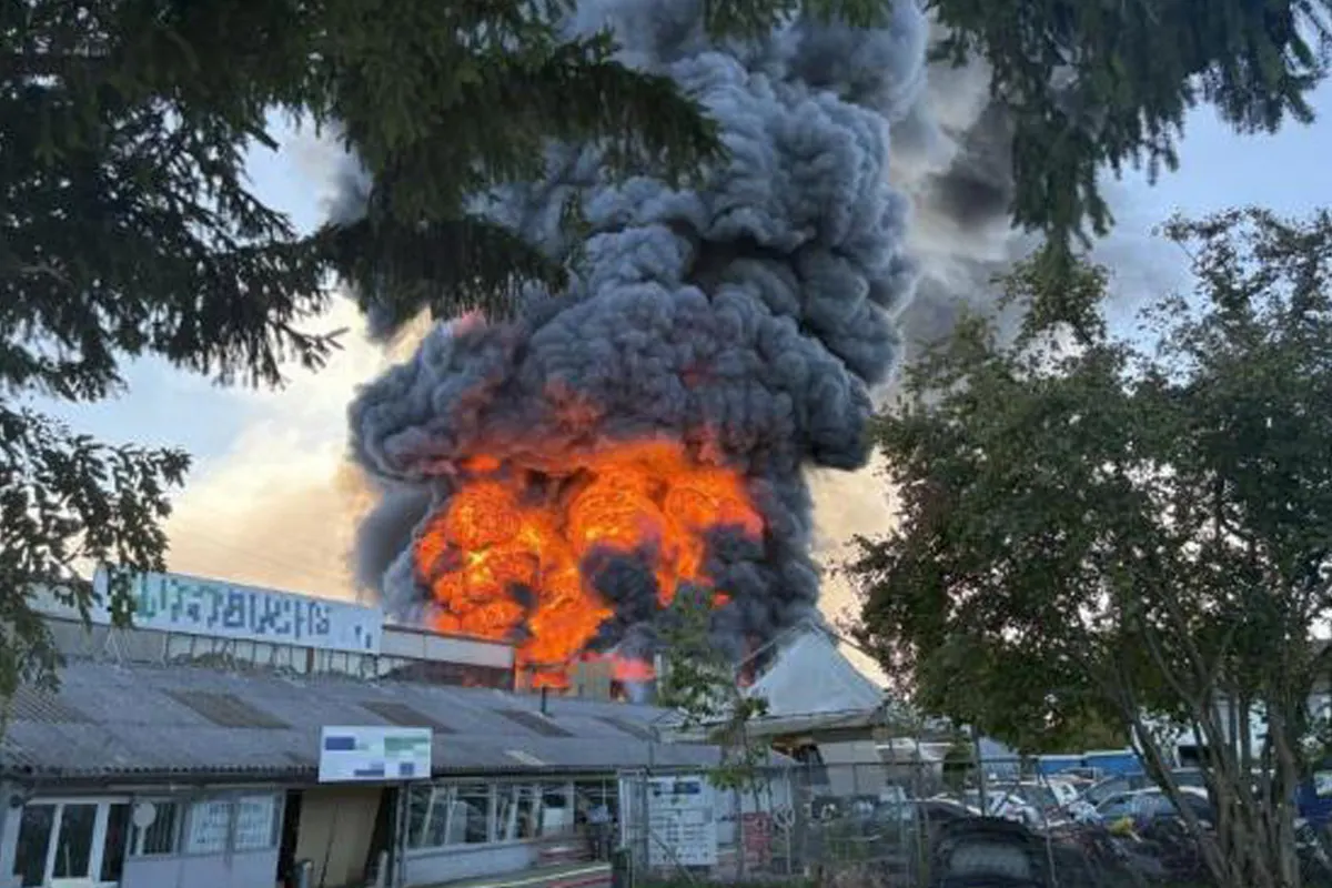 Ein Gebäudebrand mit intensiven Flammen, die durch dichte, schwarze Rauchwolken aufsteigen. Die Umgebung zeigt Bäume und ein Gebäude mit sichtbaren Flammen.