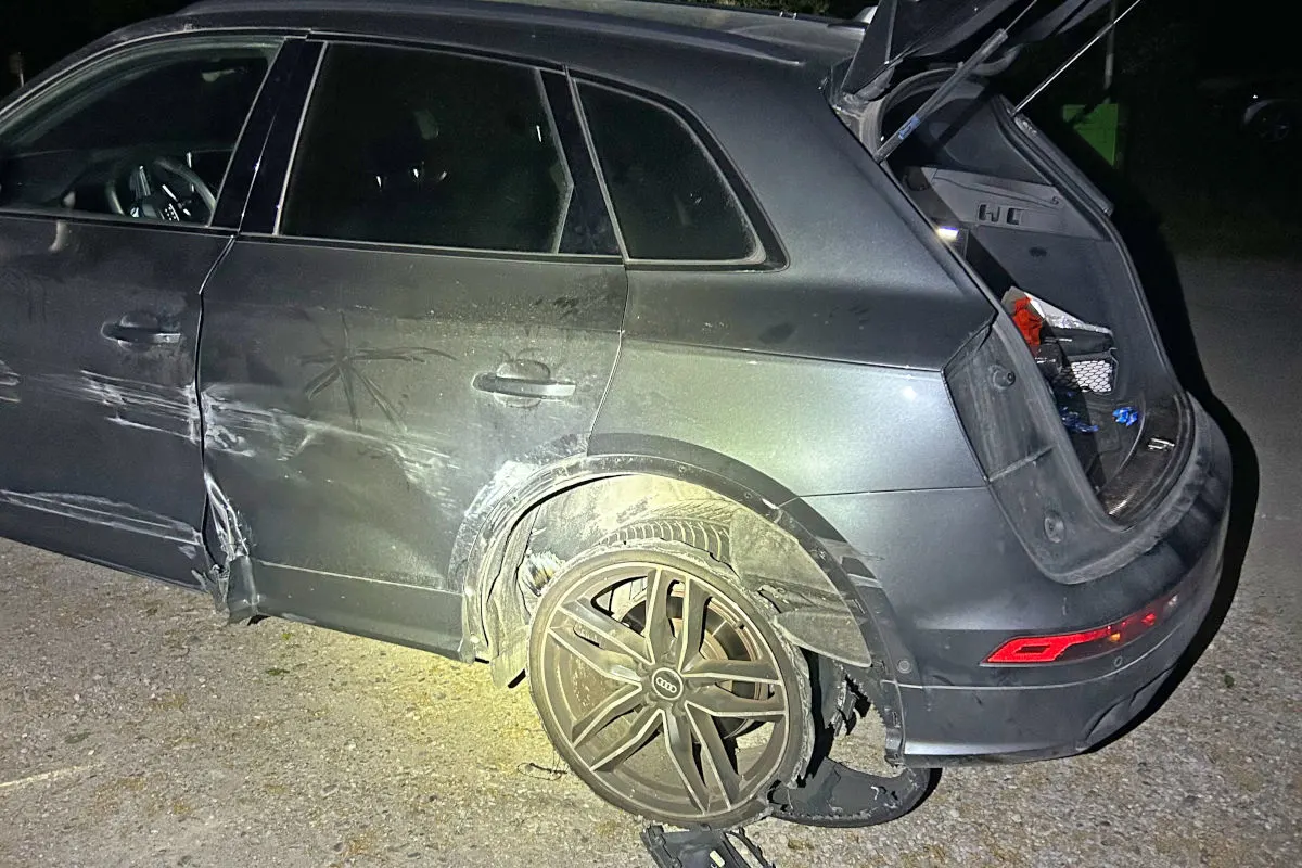 Ein graues Auto mit kaputtem Reifen und zerbeulter Seite steht bei Nacht auf einer Straße. Der Kofferraum ist offen, und der Innenraum ist teilweise sichtbar.