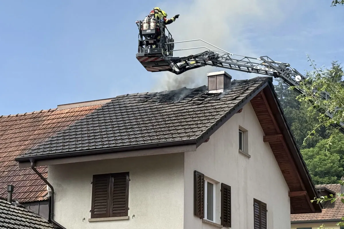 Ein Feuerwehrmann steht auf einer Drehleiter und löscht Rauch auf dem Dach eines Wohnhauses. Es ist ein sonniger Tag.